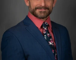 A polished studio headshot with formal business styling. Professional studio headshot of a man wearing a blue suit and patterned tie.