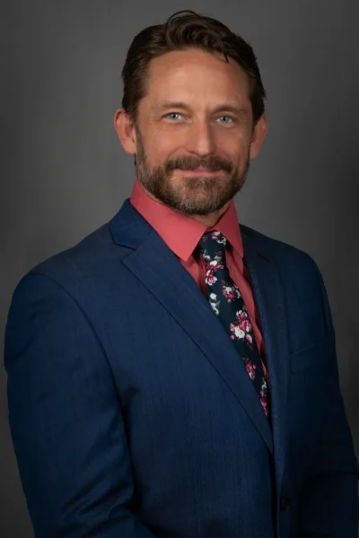 A polished studio headshot with formal business styling. Professional studio headshot of a man wearing a blue suit and patterned tie.