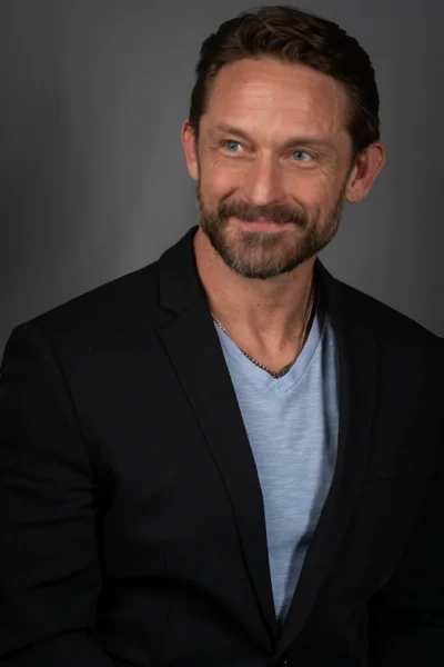 A relaxed professional portrait with clean studio lighting. Professional studio portrait of a man wearing a black jacket and blue shirt.