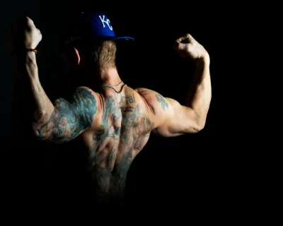 Strength, shadow, and ink. Dramatic low-key portrait of a tattooed man seen from behind, flexing his arms while wearing a Kansas City cap.