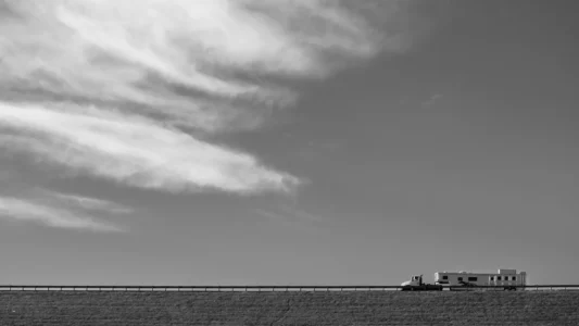 A small truck pulling a horse trailer crosses a quiet ridgeline beneath a vast, clouded sky. Black and white photograph of an truck pulling a horse trailer traveling along a distant ridgeline beneath a large open sky with sweeping clouds.