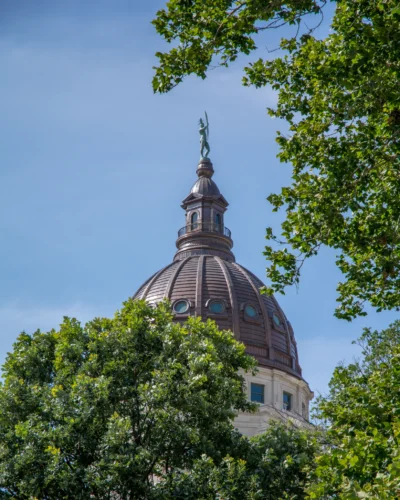 The Kansas State Capitol dome in Topeka, Kansas, seen through summer trees. Kansas State Capitol dome in Topeka, Kansas, partially framed by green trees under a blue sky.