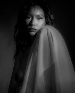 A quiet black-and-white portrait shaped by shadow, gaze, and the soft form of draped fabric. Black-and-white fine art portrait of a woman gazing at the camera with sheer fabric draped over one shoulder.