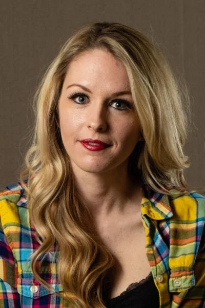 A confident editorial portrait with bold styling and direct eye contact. Editorial portrait of a blonde woman wearing a colorful plaid shirt.