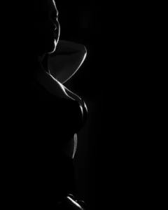 A standing figure study shaped by rim light, shadow, and quiet contour. Low-key black and white fine art image of a standing woman in partial silhouette with rim light defining her face, arm, and upper body.