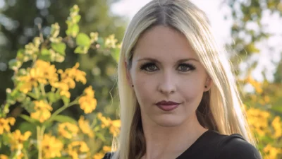 A natural-light outdoor portrait framed by soft greenery and yellow flowers. Outdoor portrait of a blonde woman with yellow flowers in the background.