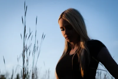 A quiet outdoor portrait with strong profile shape and open sky. Outdoor portrait of a woman in profile near tall grass against a blue sky.