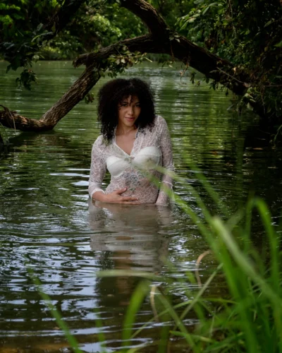A quiet portrait in water and shade. Environmental portrait of a woman in a white lace dress standing in shallow water beneath overhanging tree branches.