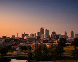 Downtown Kansas City glows in golden-hour light as the skyline rises above the surrounding roads, trees, and urban landscape. Kansas City skyline at sunset with warm orange light, downtown buildings, highways, trees, and a darkening blue sky.