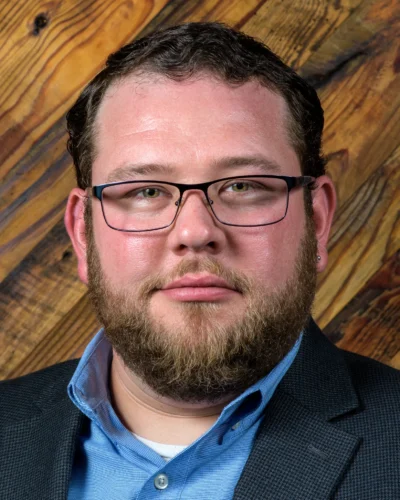 A professional headshot with a warm wood-textured background. Professional headshot of a man wearing glasses and a blazer in front of a wood background.