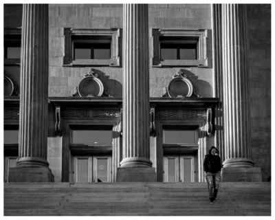 A lone figure passes through the steps and columns of the Idaho State Capitol. Black-and-white photograph of a lone figure walking near the steps and columns of the Idaho State Capitol.