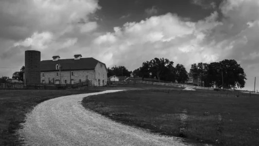A gravel road curves toward a quiet farmstead under a restless Midwestern sky. Black and white photograph of a curved gravel road leading toward a barn and silo beneath large dramatic clouds.