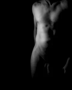 A sculptural black and white figure study emphasizing light, shadow, and the abstract form of the human torso. Black and white fine art study of a male torso emerging from deep shadow.