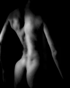 A low-key black and white figure study exploring the contours of the back, shoulders, and body through shadow and light. Black and white fine art study of a male back and figure in dramatic side lighting.