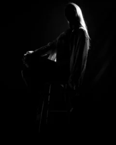 A seated silhouette study defined by rim light, posture, and darkness. Low-key black and white fine art portrait of a seated woman in silhouette with rim light on her hair and clothing.